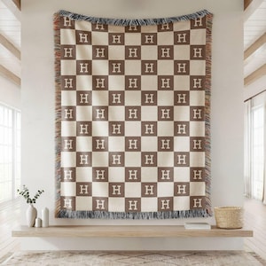 May include: A woven throw blanket featuring a checkered pattern of alternating beige and brown squares. Each square contains a white letter H. The blanket has a fringed edge with multicolored threads along the top and bottom borders.