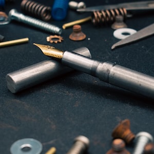 May include: A silver fountain pen with a gold nib and cap, resting on a dark surface. The pen is surrounded by various metal hardware items, including screws, springs, and washers, suggesting a mechanical or industrial theme.