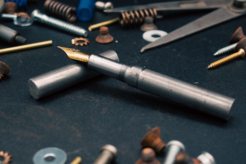 May include: A silver fountain pen with a gold nib and cap, resting on a dark surface. The pen is surrounded by various metal hardware items, including screws, springs, and washers, suggesting a mechanical or industrial theme.