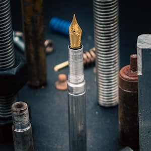 May include: A silver fountain pen with a gold nib is positioned amongst various metal bolts and hardware. The pen is the central focus, with a detailed nib and a sleek, metallic body. The background includes rusty and new metal components.
