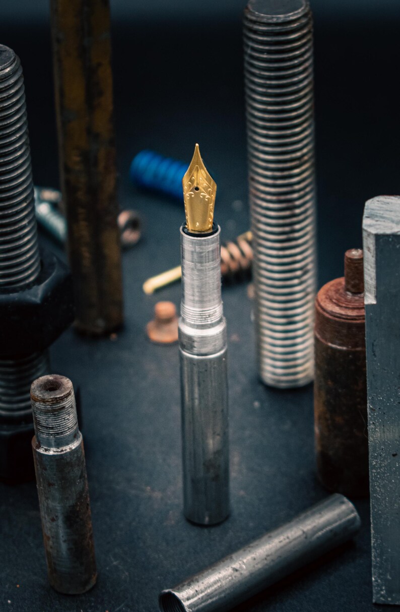 May include: A silver fountain pen with a gold nib is positioned amongst various metal bolts and hardware. The pen is the central focus, with a detailed nib and a sleek, metallic body. The background includes rusty and new metal components.