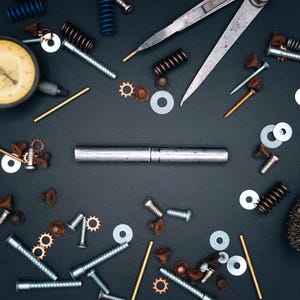 May include: A collection of metal hardware and tools, including screws, washers, springs, and a dial indicator, arranged on a dark surface. The image showcases various mechanical components, ideal for engineering or repair.