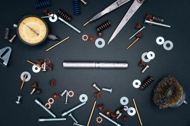 May include: A collection of metal hardware and tools, including screws, washers, springs, and a dial indicator, arranged on a dark surface. The image showcases various mechanical components, ideal for engineering or repair.