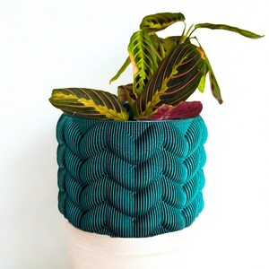 May include: A teal, textured planter with a unique, layered design, holding a houseplant with green and pink striped leaves. The planter sits atop a white base, creating a striking contrast against a white background.