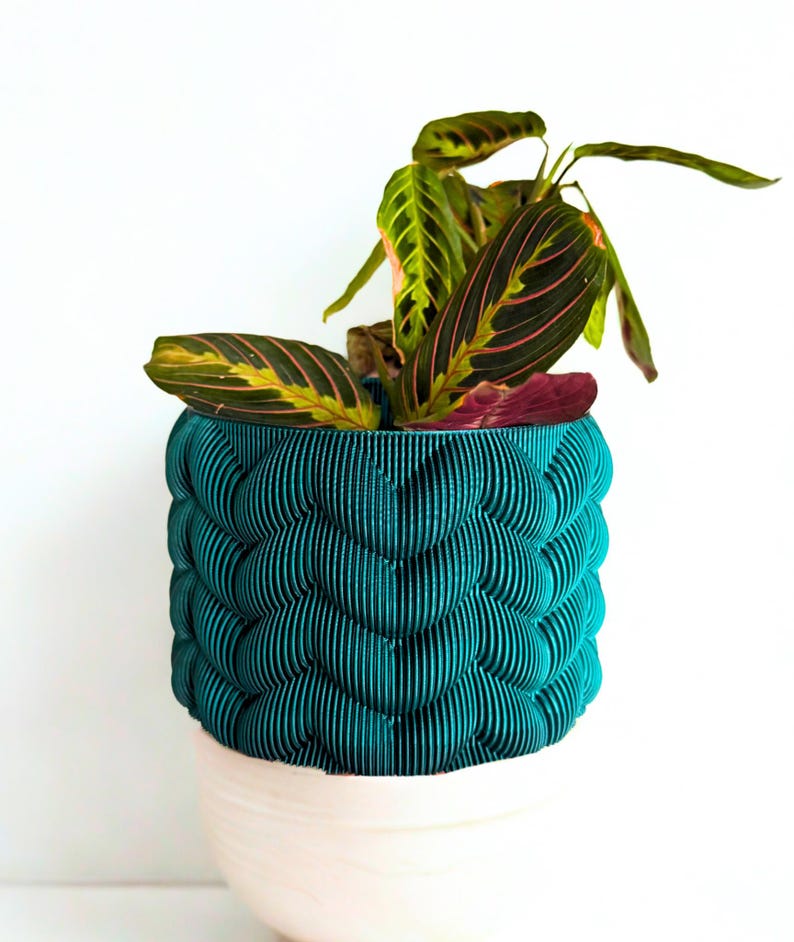 May include: A teal, textured planter with a unique, layered design, holding a houseplant with green and pink striped leaves. The planter sits atop a white base, creating a striking contrast against a white background.
