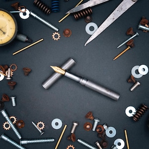 May include: A collection of metal tools and components, including a silver fountain pen with a gold nib, screws, washers, springs, and a dial gauge, arranged on a dark surface.
