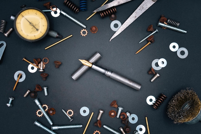 May include: A collection of metal tools and components, including a silver fountain pen with a gold nib, screws, washers, springs, and a dial gauge, arranged on a dark surface.