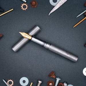 May include: A silver fountain pen with a gold nib, on a dark grey surface, surrounded by screws, washers, and gears. The pen's cap is placed beside it. The items are scattered around the pen. The image has a mechanical theme.