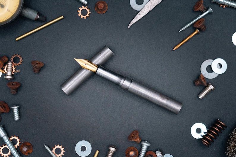 May include: A silver fountain pen with a gold nib, on a dark grey surface, surrounded by screws, washers, and gears. The pen's cap is placed beside it. The items are scattered around the pen. The image has a mechanical theme.