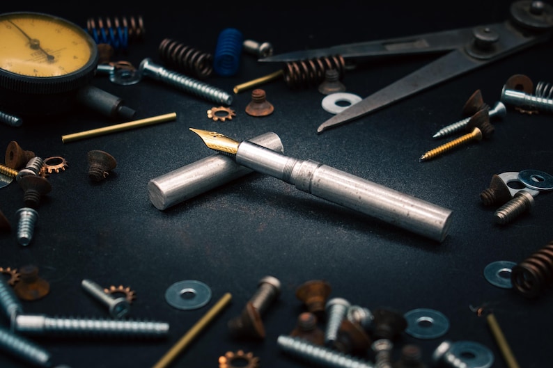 May include: A silver fountain pen with a gold nib, resting on a dark surface, surrounded by mechanical parts such as screws, springs, and washers. A yellow dial gauge is visible in the background.