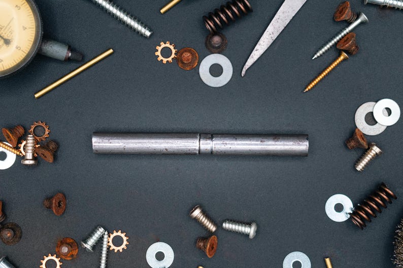May include: A collection of metal engineering components, such as screws, washers, springs, and gears, displayed on a dark background. A central metal rod is bisected. A dial gauge and a metal ruler are also present, suggesting a mechanical theme.