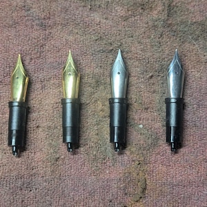 May include: Four fountain pen nibs are shown, two gold and two silver. Each nib is connected to a black section. The nibs are arranged horizontally against a textured, brown background. The gold nibs have a shiny, polished appearance.