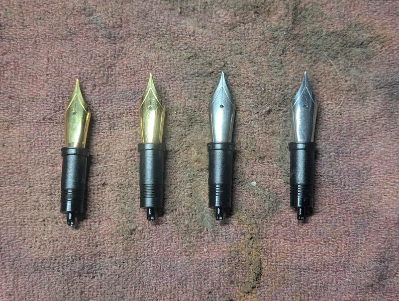 May include: Four fountain pen nibs are shown, two gold and two silver. Each nib is connected to a black section. The nibs are arranged horizontally against a textured, brown background. The gold nibs have a shiny, polished appearance.