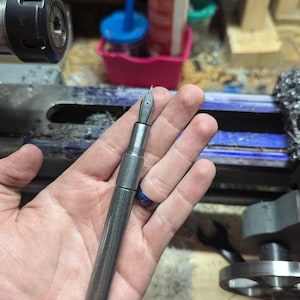 May include: A silver fountain pen with a nib, held in a hand. The pen has a cylindrical body and a cap. The background includes workshop tools and equipment, suggesting a handcrafted item.