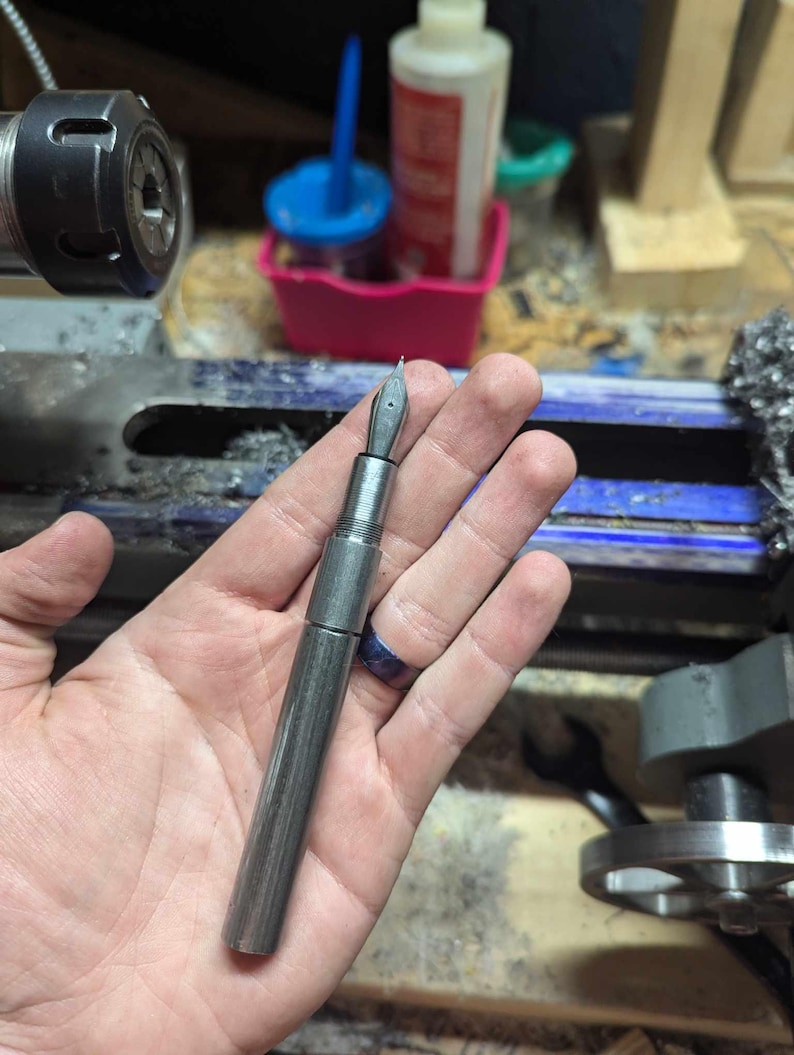 May include: A silver fountain pen with a nib, held in a hand. The pen has a cylindrical body and a cap. The background includes workshop tools and equipment, suggesting a handcrafted item.