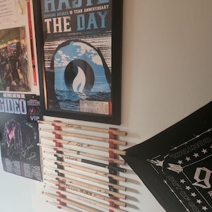 May include: A wall mounted drum stick holder with red and black accents. The holder is filled with wooden drum sticks. A black and white bandana with white stars and the text "Out of Control" is hanging on the wall. A framed poster with the text "Haste The Day" is hanging on the wall.
