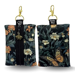 Puede incluir: Dos pequeñas bolsas rectangulares con herrajes dorados. Una bolsa tiene una cremallera negra y un dije de abeja. Ambas bolsas tienen un patrón floral oscuro con flores naranjas y blancas. La otra bolsa tiene un diseño de mariposa y el texto "Sweetie Bird".