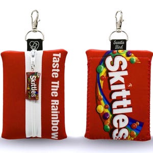 May include: Red Skittles-themed pouch with a white zip and a small Skittles candy pack attached. The pouch has the text "Taste The Rainbow" on the side. A silver clasp and a black tag with a white logo are attached to the top.