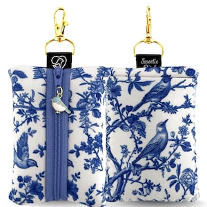 May include: A rectangular fabric pouch featuring a blue and white toile pattern of birds and branches. It includes a blue zipper, a gold-tone lobster clasp, and a small bird-shaped charm. One side shows a black tag with a white bird icon.