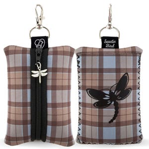 Może przedstawiać: Dwie materiałowe saszetki w brązowo-niebiesko-beżową kratę ze srebrnym klipsem. Jedna ma czarny zamek błyskawiczny i zawieszkę w kształcie ważki. Druga ma czarną grafikę ważki. Obie mają tekst.