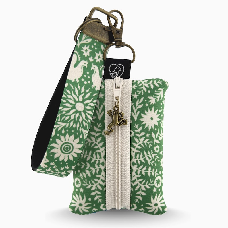 May include: Green floral print pouch with a cream zipper and a frog charm. The pouch has a matching wrist strap with a white and green pattern. The metal hardware is bronze-colored.