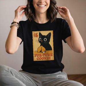 Könnte beinhalten: Schwarzes T-Shirt mit einer Cartoon-Katze und einem Glückskeks-Motiv. Das Design enthält den Text "FUR-TUNE COOKIE" und ein chinesisches Schriftzeichen. Die Grafik befindet sich auf einem beigefarbenen Hintergrund.