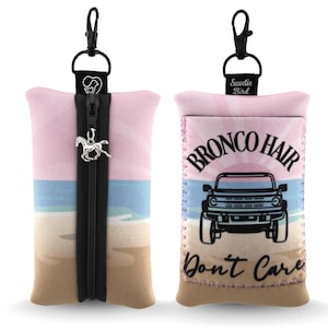 Puede incluir: Un estuche rectangular con cremallera negra y un dije de caballo plateado. La tela muestra una escena de playa con cielo rosa, agua azul y arena beige. Un lado presenta un gráfico negro de un camión con el texto Bronco Hair Don't Care.