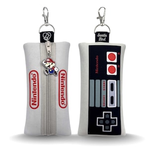 May include: Two white rectangular pouches with silver zippers and keychains. One features the Nintendo logo in red, the other a black and gray controller design. A Mario charm hangs from the zipper.