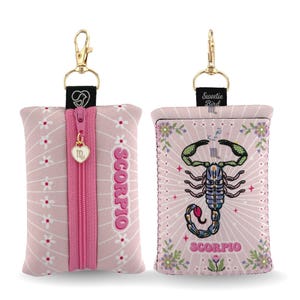 Pode incluir: Bolsa de tecido rosa com padrão floral e zíper rosa. A bolsa tem um clipe dourado e um pingente em forma de coração. A palavra "Scorpio" é bordada na lateral. O outro lado apresenta uma ilustração de escorpião com a palavra "Scorpio".