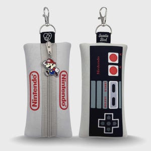 May include: Two gray Nintendo-themed pouches with silver hardware. One pouch features a Mario charm on the zipper and the Nintendo logo. The other pouch has a controller design with the Nintendo logo and the text "Sweetie Bird".