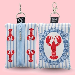 Puede incluir: Dos pequeñas bolsas rectangulares con diseños de langosta. Una bolsa tiene un patrón de rayas azules y blancas con gráficos de langosta roja y una cremallera. La otra bolsa tiene un gráfico de langosta roja dentro de un marco adornado azul y blanco. Ambas tienen un cierre plateado.