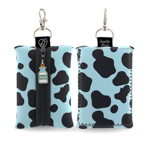 May include: A light blue neoprene keychain holder with a black cow print. The holder has a black zipper with a milk bottle charm and a silver clasp. The top of the holder has a black tag with a silver clasp and a black tag with the text "Sweetie Bird".