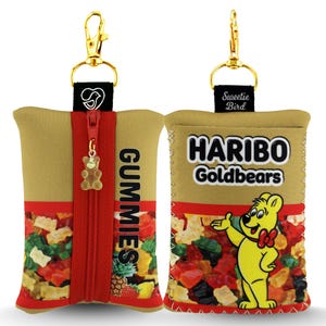 Può includere: Un astuccio beige e rosso con cerniera e clip dorata, con un ciondolo a forma di orsetto gommoso. Sull'astuccio è stampata la scritta "GUMMIES". L'altro lato presenta il logo Haribo Goldbears.