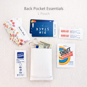 May include: A white L pouch with the text "Back Pocket Essentials" and "L Pouch". The pouch contains scissors, cash, and a blue card. Other items include lens cleaning wipes, Band-Aids, and a Shout wipe.