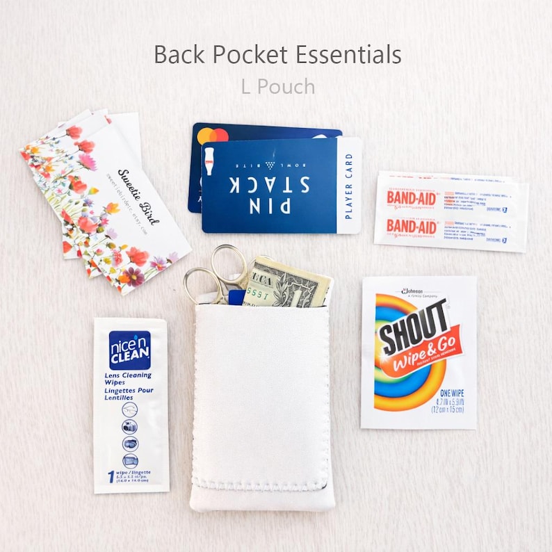 May include: A white L pouch with the text "Back Pocket Essentials" and "L Pouch". The pouch contains scissors, cash, and a blue card. Other items include lens cleaning wipes, Band-Aids, and a Shout wipe.