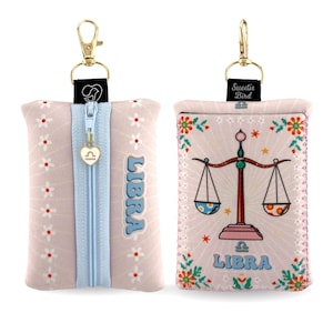 May include: A rectangular pouch with a light pink background featuring white daisy patterns and the word LIBRA. It includes a light blue zipper with a heart-shaped charm, a gold lobster clasp, and a graphic of a scale on the reverse side.