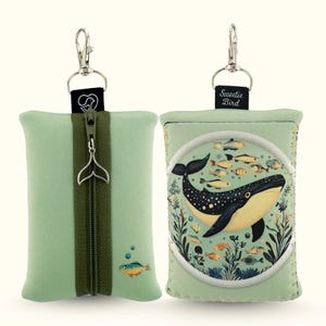 May include: Two light green rectangular pouches with metal lobster clasps. One features a central olive green zipper with a whale tail charm and a small fish graphic. The other displays a circular illustration of a whale surrounded by smaller fish.