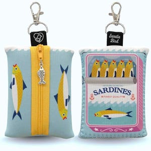 May include: A light blue zippered pouch with a yellow zipper and fish charm. The pouch features a fish pattern on one side and a sardine can design on the other. The text "Sardines Finest Quality" is visible.