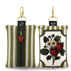 May include: Two small, rectangular pouches with gold-colored hardware. One pouch has vertical olive green and white stripes, a black zipper, and a strawberry charm. The other pouch features a floral strawberry design with the text "Sweetie Bird".