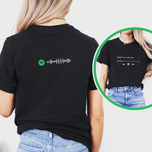 May include: Black t-shirt featuring a Spotify code on the back and a music player graphic with the text "I Want You To Want Me" on the front. The music player graphic includes play, pause, and skip buttons.