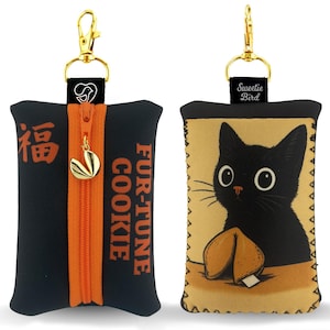 Op de afbeelding: Zwarte en oranje fortune cookie-vormige etui met een gouden rits en clip. Eén kant heeft Chinese karakters en de woorden "FUR-TUNE COOKIE". De andere kant heeft een illustratie van een zwarte kat met een fortune cookie.