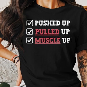 Puede incluir: Camiseta negra con una marca de verificación blanca junto a las palabras "PUSHED UP", "PULLED UP" y "MUSCLE UP". Las palabras "MUSCLE UP" están en rojo.