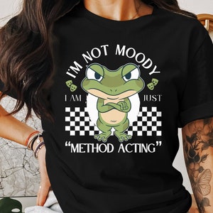 Könnte beinhalten: Schwarzes T-Shirt mit einer grünen Frosch-Karikatur und dem Text "I'm not moody I am just "method acting""