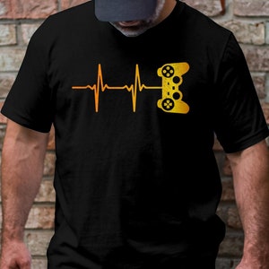 Puede incluir: Una camiseta negra con un gráfico amarillo de un mando de videojuego y una línea de latido del corazón.