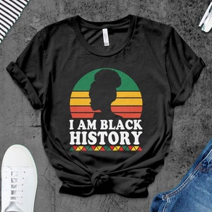 May include: Black t-shirt featuring a retro sunset graphic and the text "I AM BLACK HISTORY" in white. The graphic depicts a silhouette of a woman with an afro.