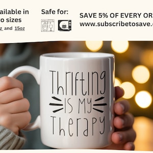 May include: White ceramic mug with black text that reads "Thrifting is my therapy". The mug is being held by a hand against a blurry background of warm yellow lights.