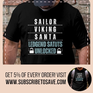 May include: Black t-shirt with white text that reads "Sailor Viking Santa Legend Status Unlocked" with a padlock icon on either side of the word "Unlocked".