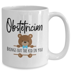 May include: White ceramic coffee mug with a black and brown illustration of a teddy bear wearing a blue bow tie. The text "Obstetrician brings out the kid in you" is printed on the mug.
