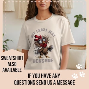 May include: Cream-colored t-shirt featuring a cartoon chicken with the text "NOT CRAZY JUST HENSANE". The chicken has red comb and wattle, with a brown and gray body. The shirt is a casual wear item.