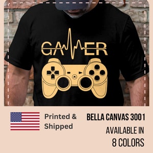 Puede incluir: Camiseta negra con un gráfico dorado de un mando de videojuego y la palabra "GAMER" con un gráfico de latido del corazón. Impreso y enviado. Bella Canvas 3001. Disponible en 8 colores.